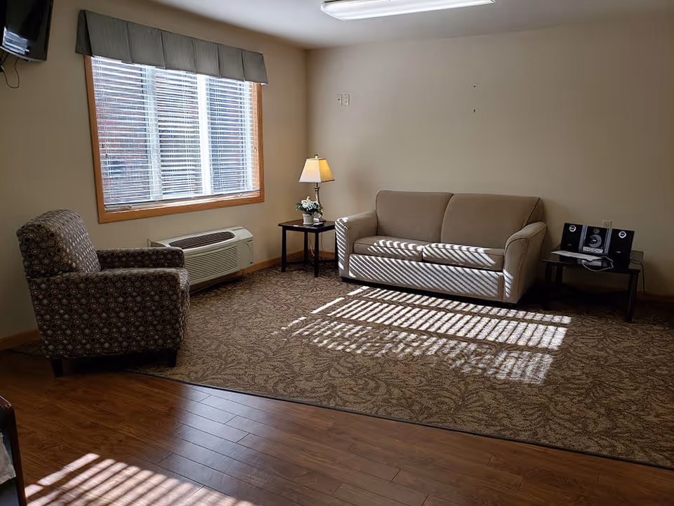 Cozy senior living lounge with sofa, armchair, side tables, lamp, window blinds, and sunlight.