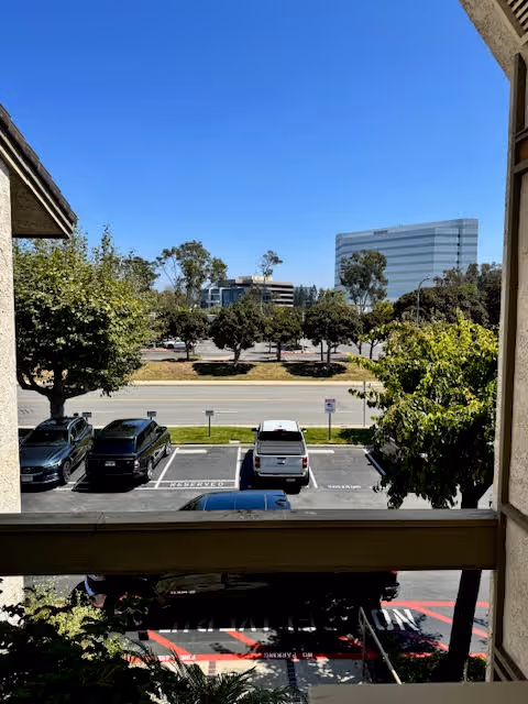 Balcony view overlooking a parking lot, trees, and a distant office building beneath blue sky.