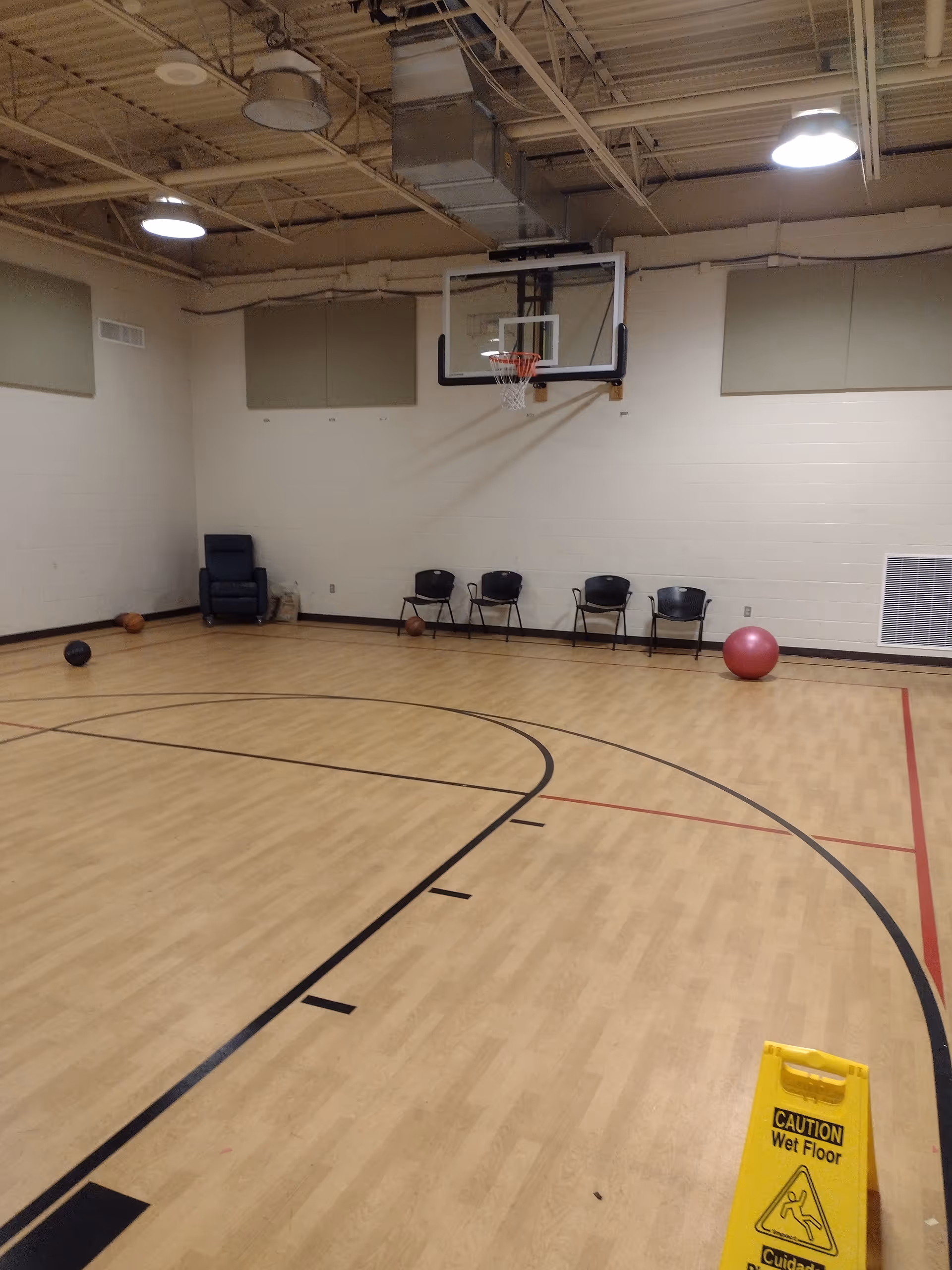 Indoor gym with basketball hoop, wooden court, chairs along wall, and wet floor sign.