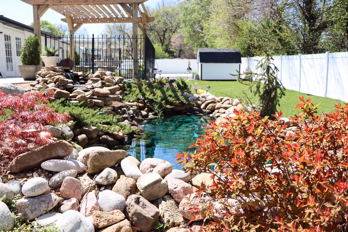 Outdoor landscaped garden with a rocky pond, waterfall, pergola, and fence in background.