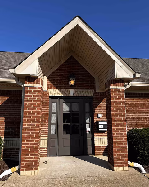 Brick building entrance with a gabled, peaked roof overhang and double doors.