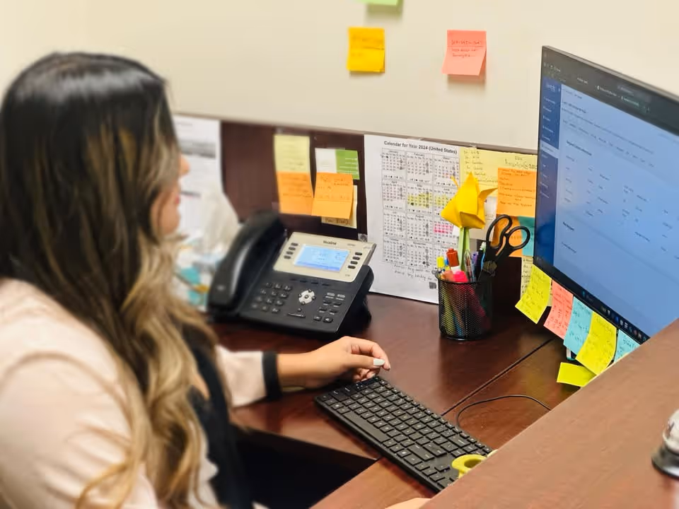 Office workspace with a staff member at a desk, computer, phone, and colorful sticky notes.