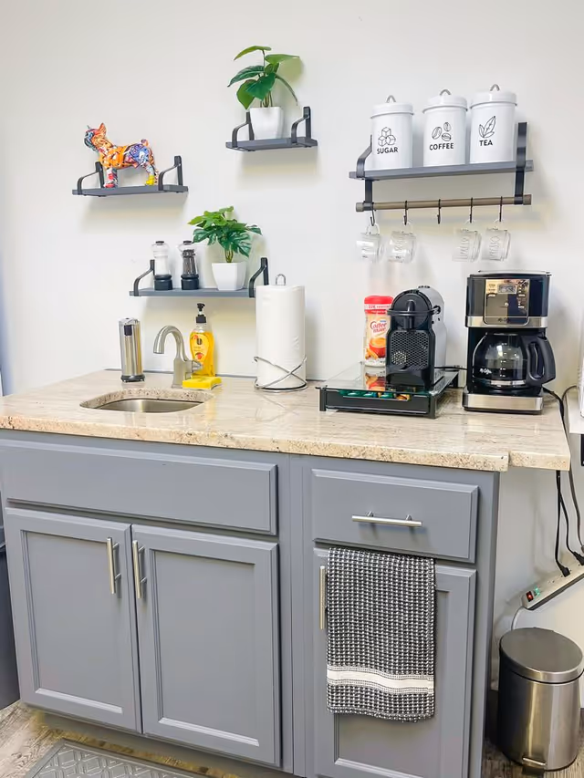 Modern kitchenette with gray cabinets, coffee station, shelves, and potted plants.