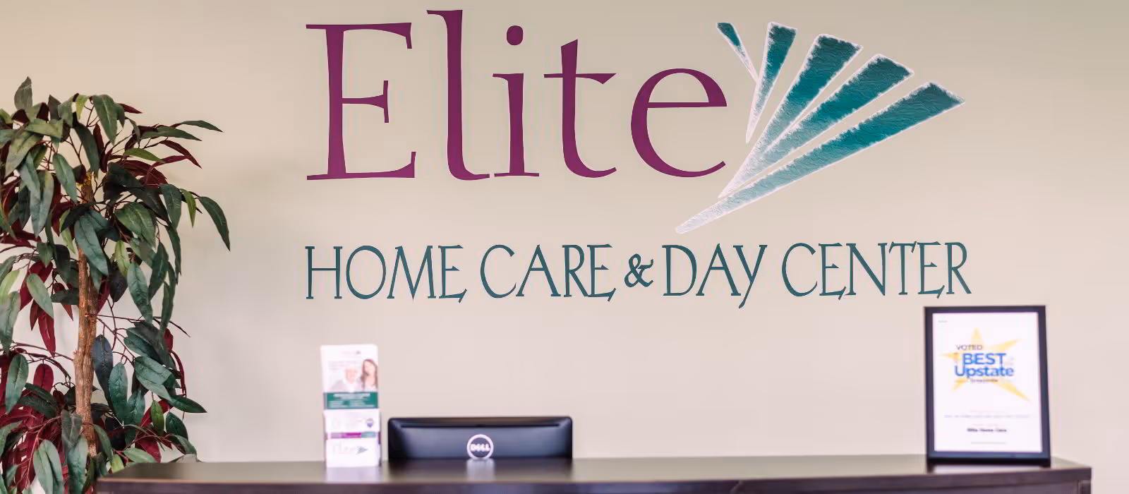 Lobby with reception desk, computer, brochure stand, plant, and Elite Home Care & Day Center sign.
