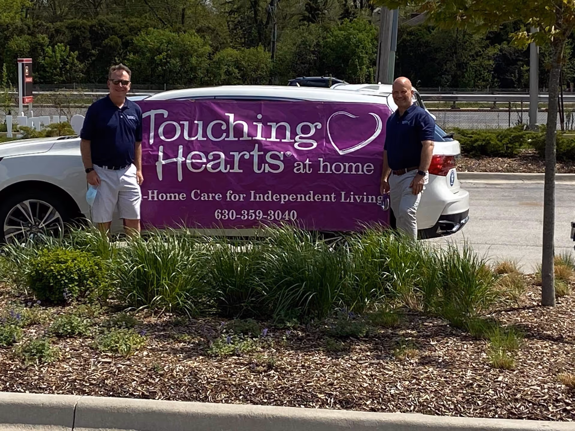 Two men stand beside a white car with a large purple Touching Hearts at Home banner.