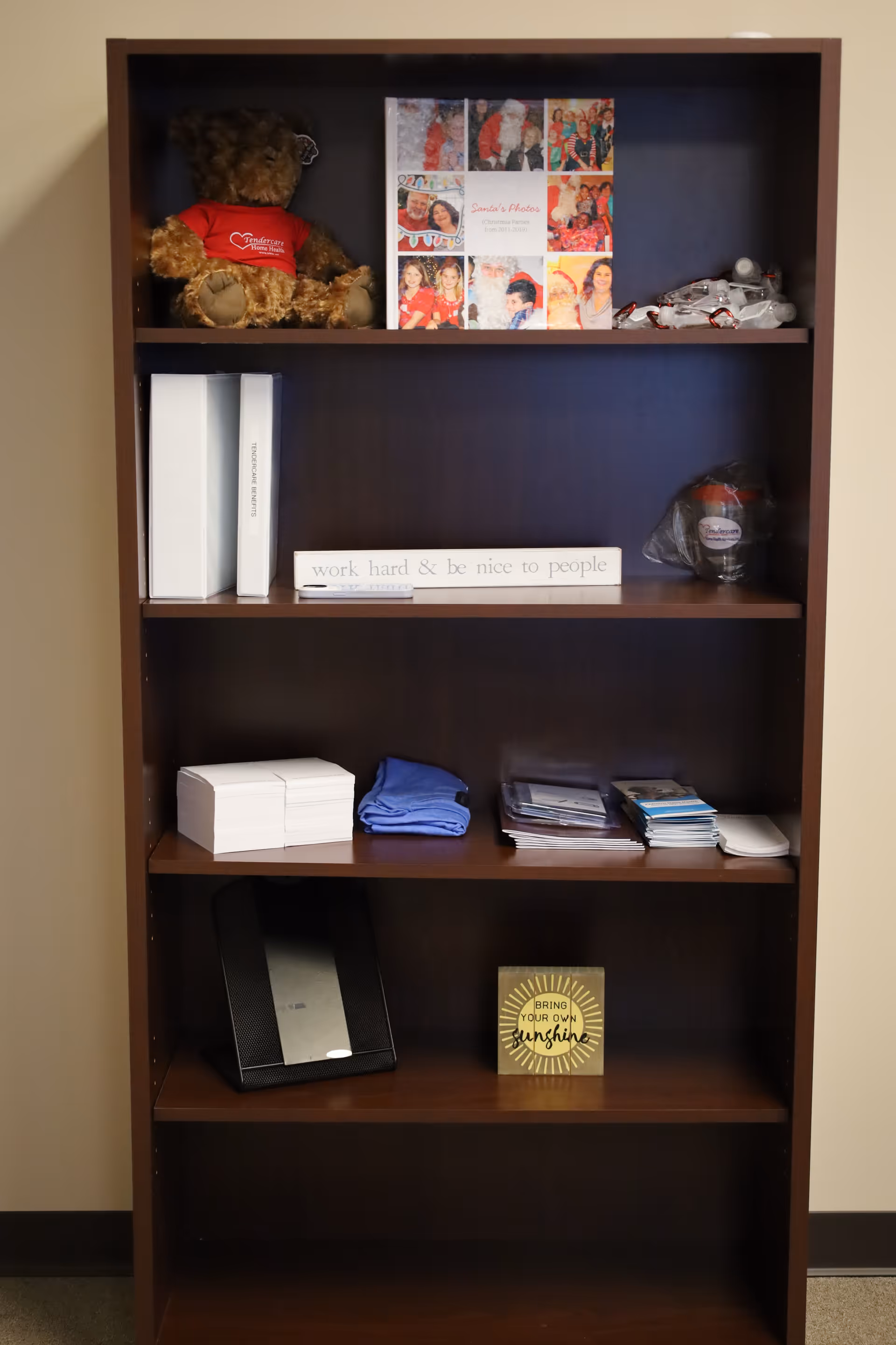 Tall wooden bookshelf in a hallway holds a teddy bear, photos, and assorted papers.