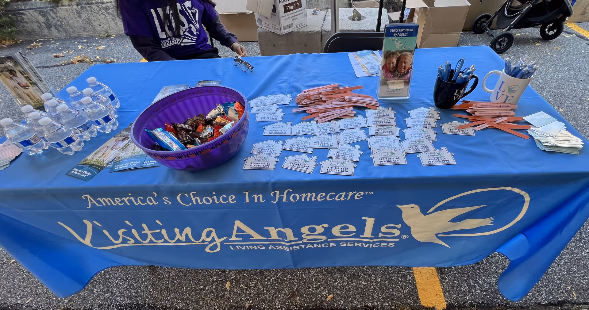Promotional table with Visiting Angels branding, flyers, pens, candy, and bottled water outdoors.
