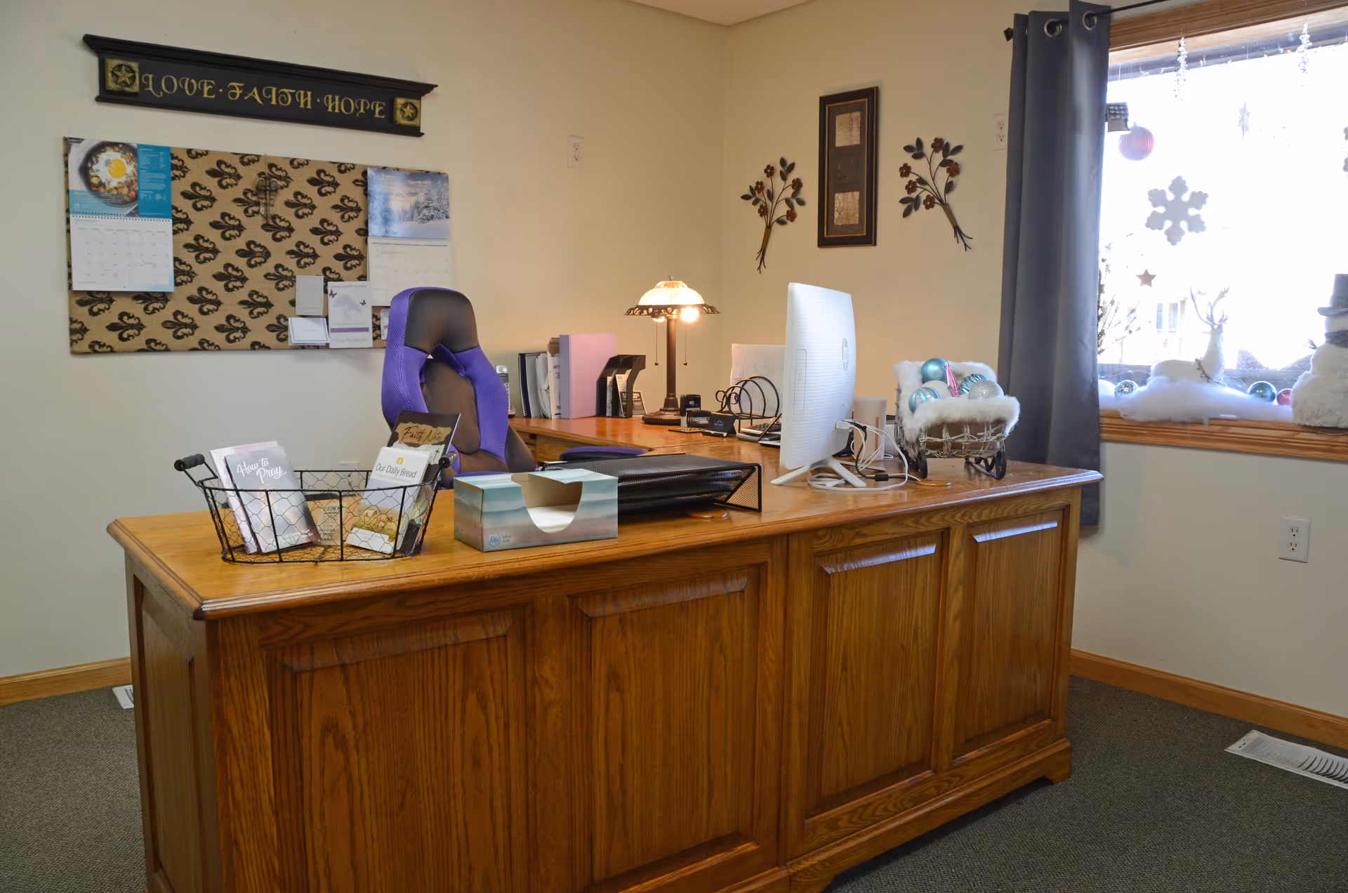 Wooden reception desk with computer, calendar, lamp, and winter-themed window decorations.