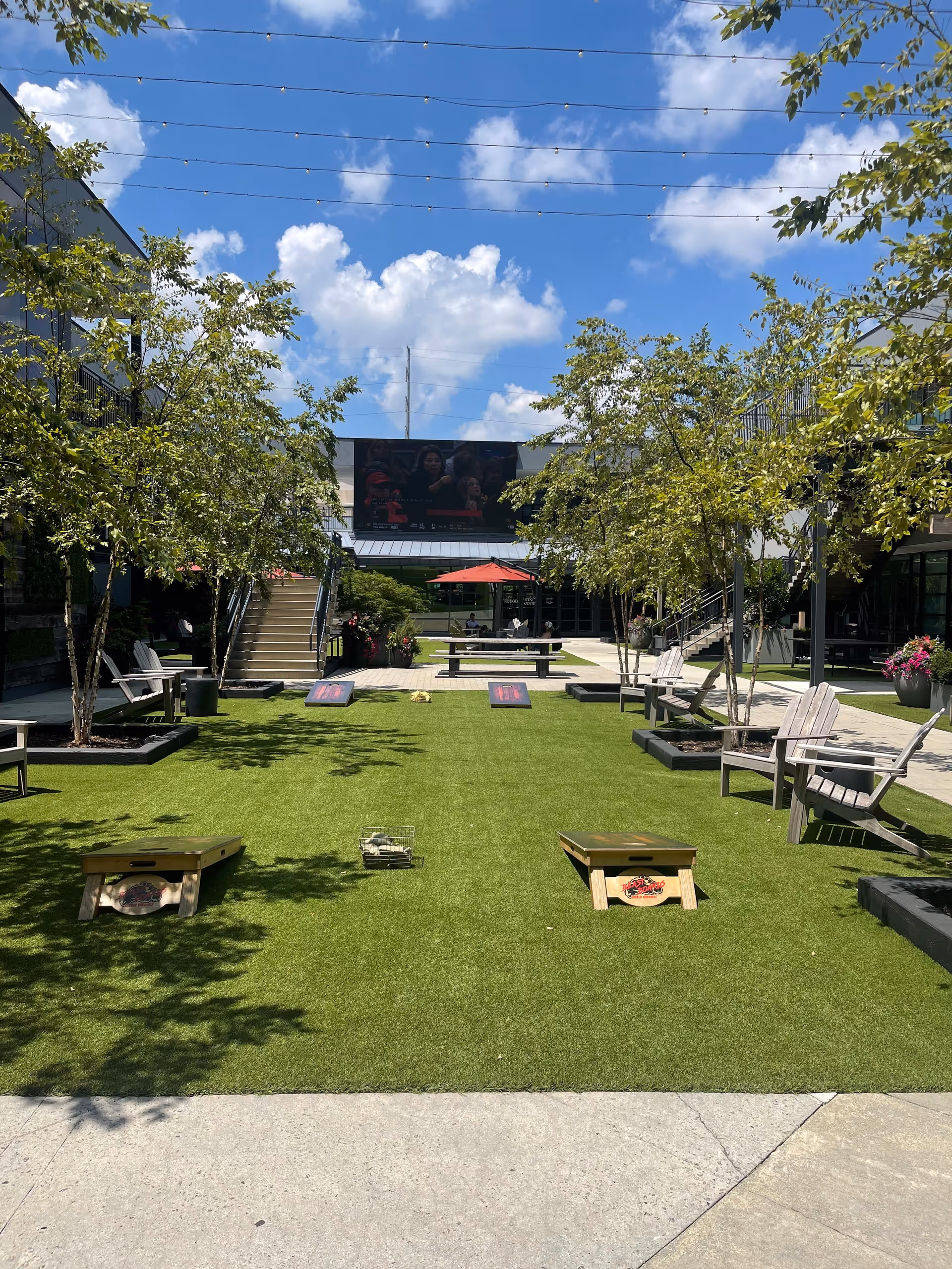 Outdoor courtyard with artificial turf, cornhole boards, trees, and seating.