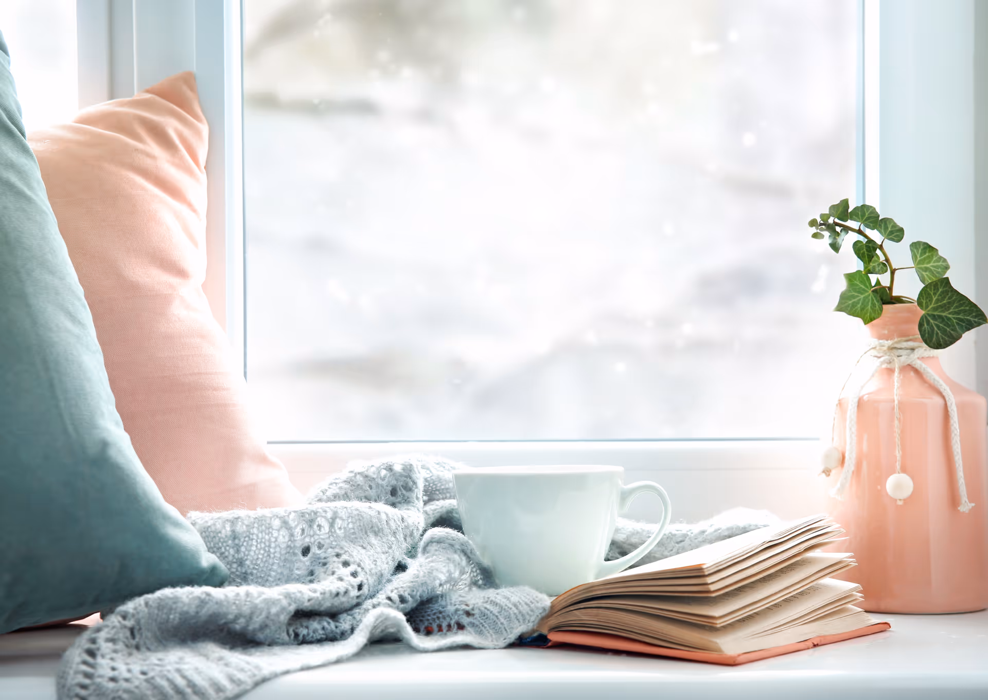 Cozy windowsill scene with pastel cushions, a knitted blanket, mug, open book, and potted plant.