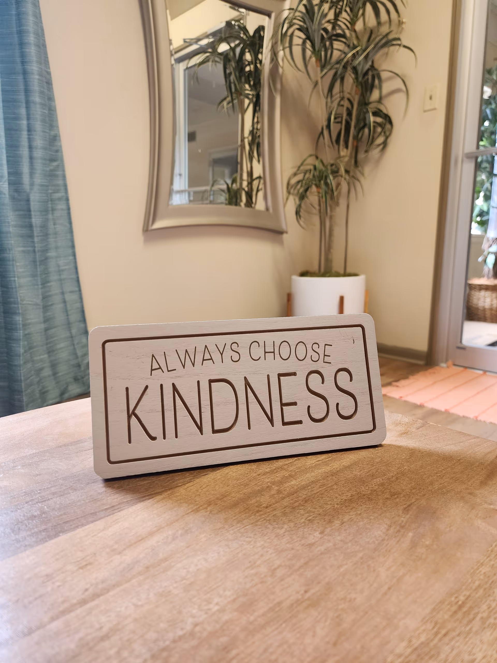 A wooden table holds a decorative Always Choose Kindness sign inside a warm interior.