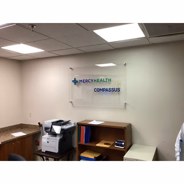 Office reception area with printer, shelves, and Mercy Health Compassus sign on wall.
