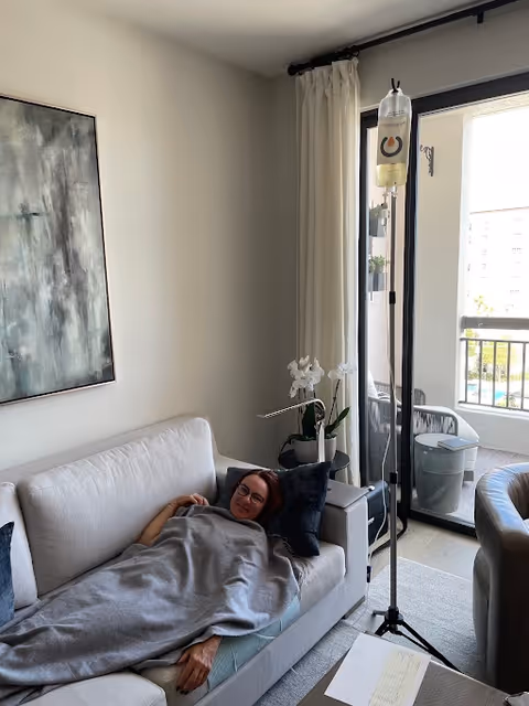 Senior resident resting on a gray sofa with an IV drip stand by a balcony door.