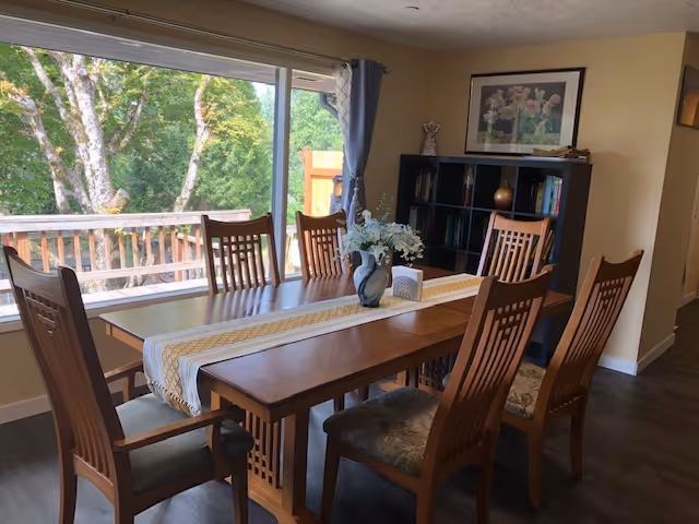 A well-lit dining room with a long wooden table, eight chairs, and large window view.