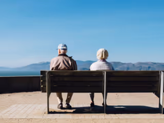 Two seniors sit on a bench overlooking distant mountains outdoors.