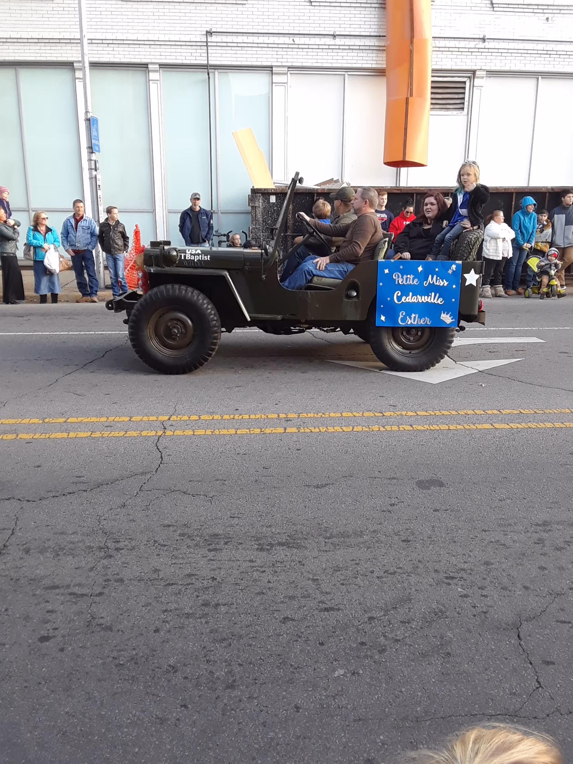 A vintage jeep with riders drives past a crowd on a city street parade