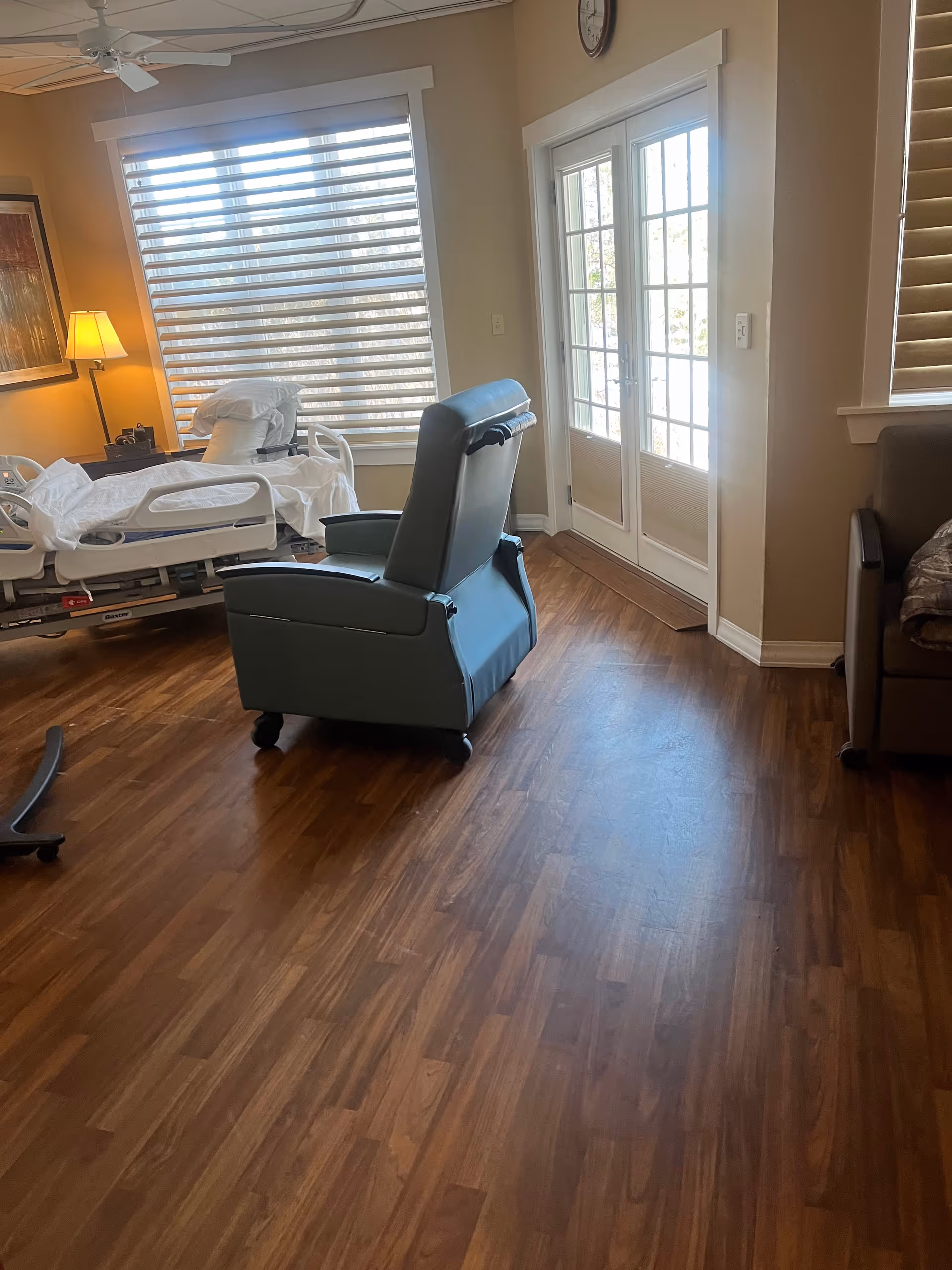 Interior patient room with a hospital bed, blue recliner, blinds, and glass door.