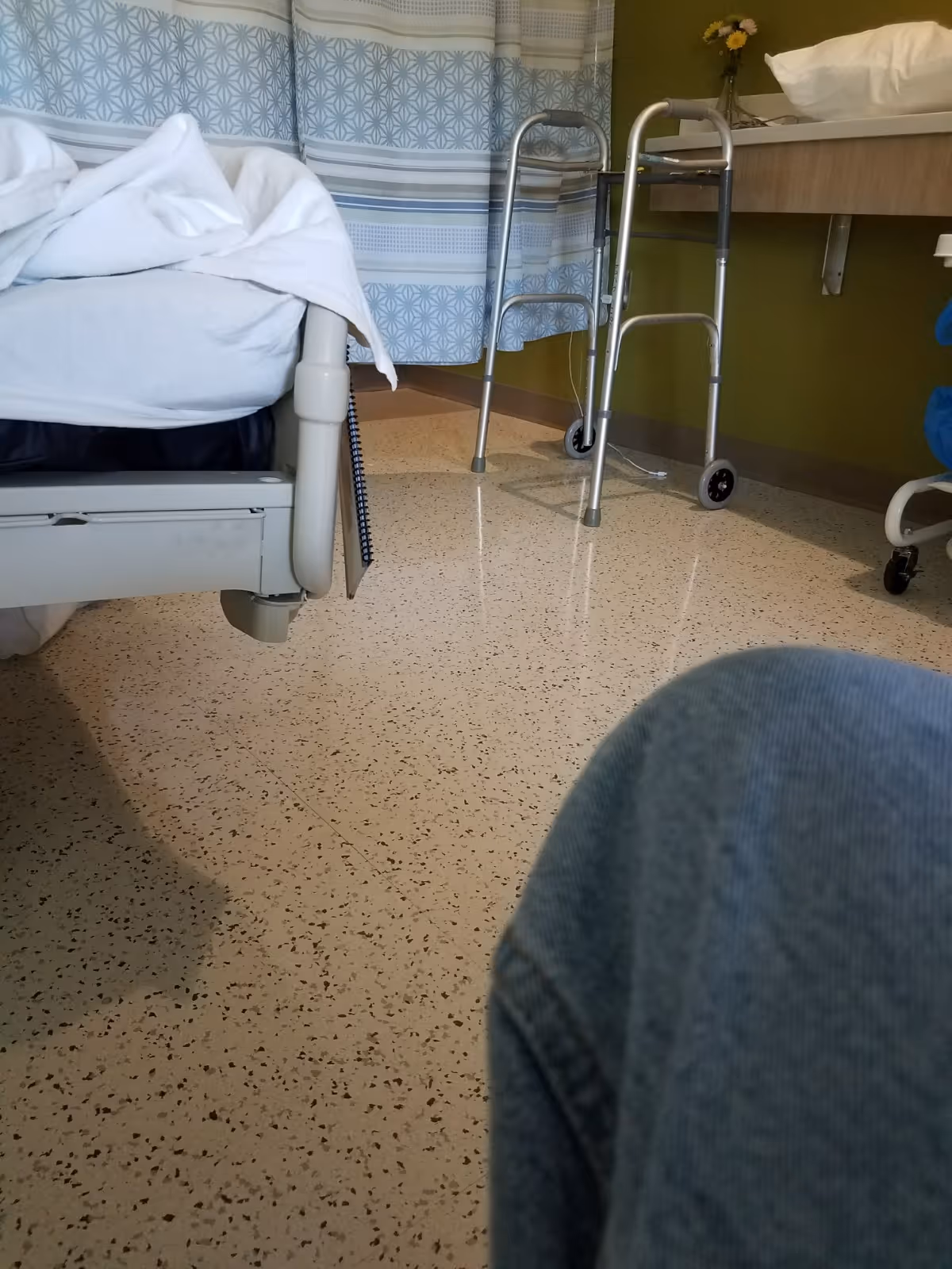 Hospital patient room with bed, privacy curtain, walker, and patterned flooring.