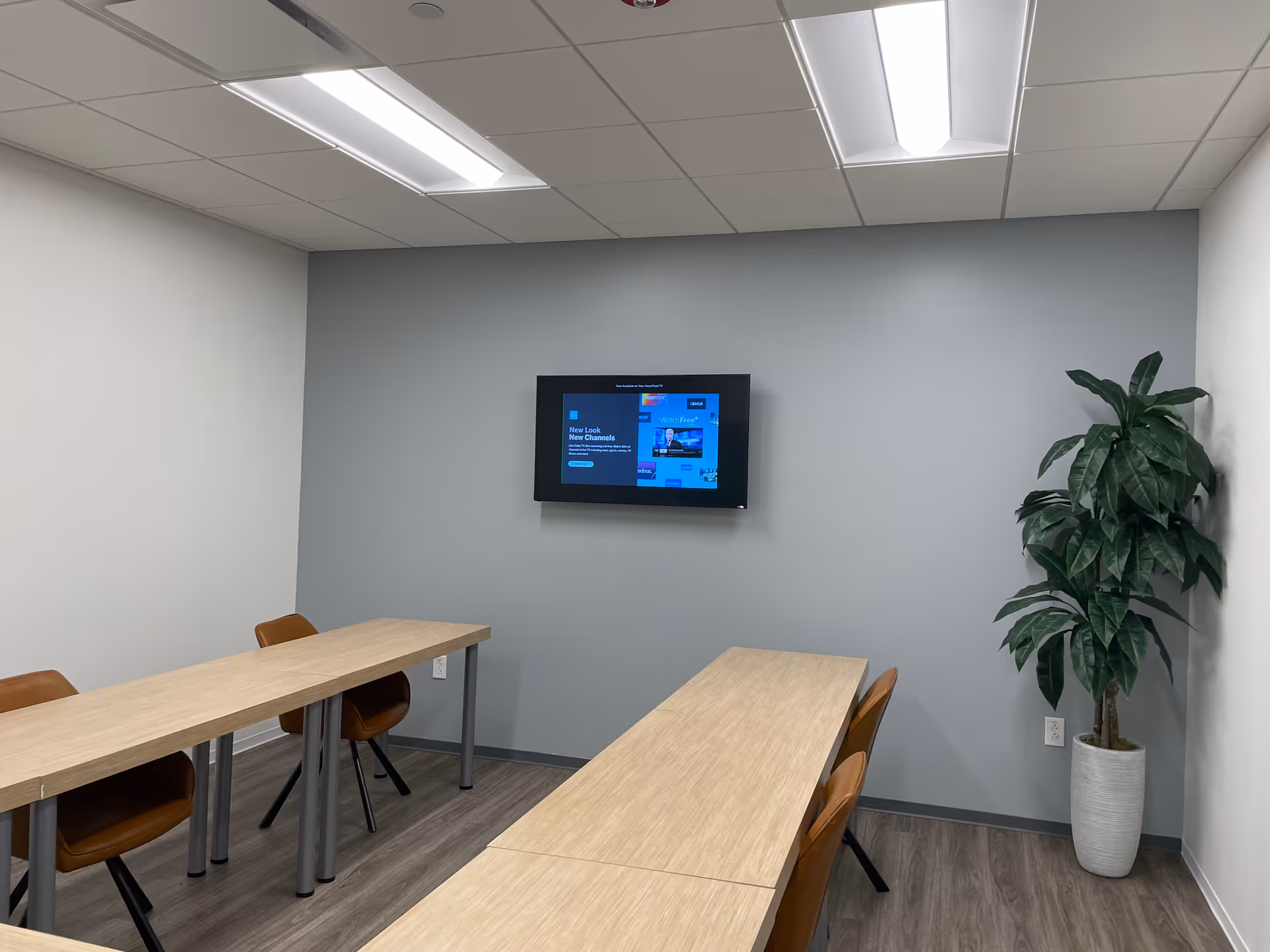 A modern meeting room with L-shaped desks, chairs, a wall-mounted TV, and a potted plant.