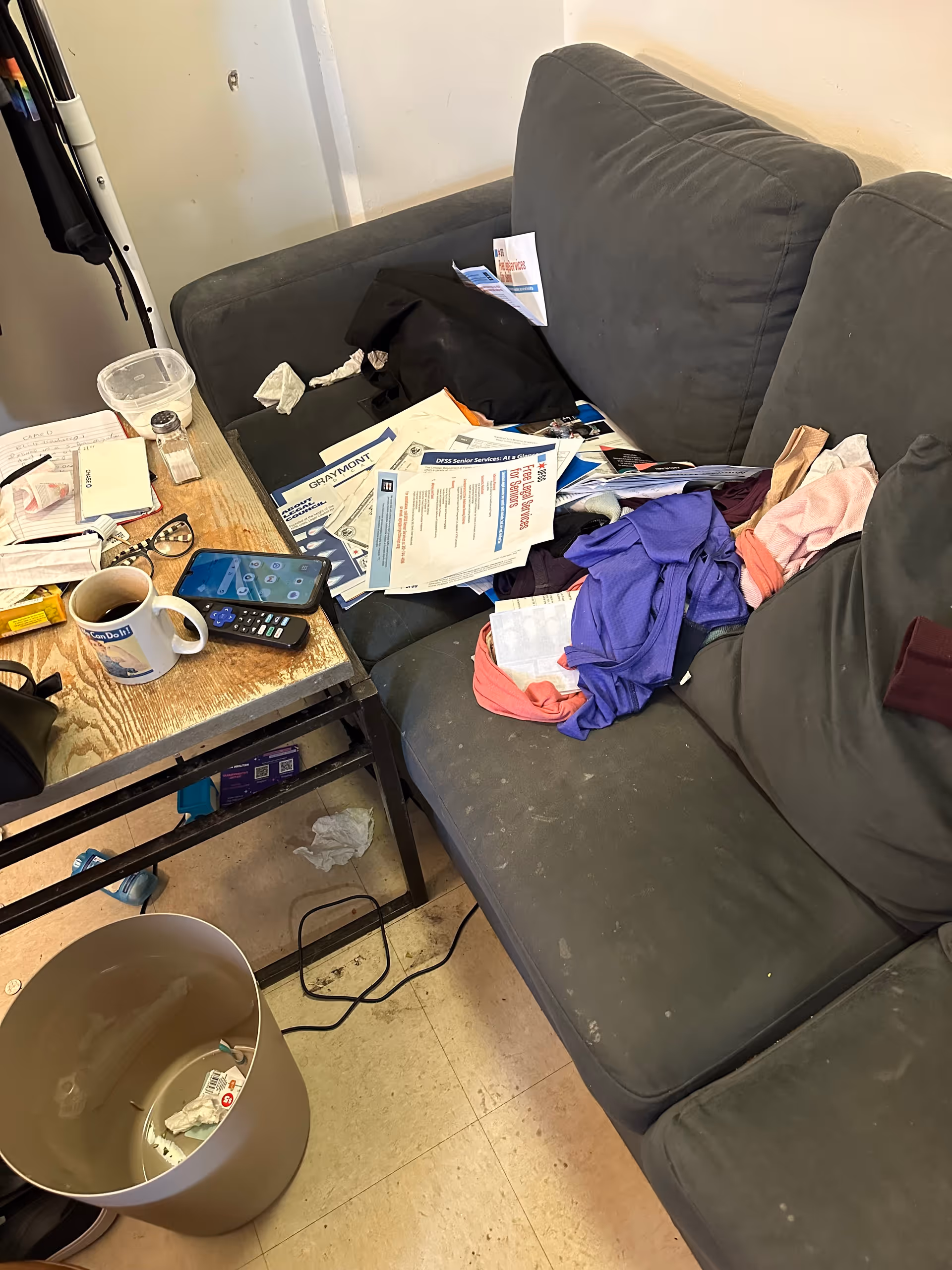 Messy living room with a sofa, cluttered table, papers, mug, and trash.