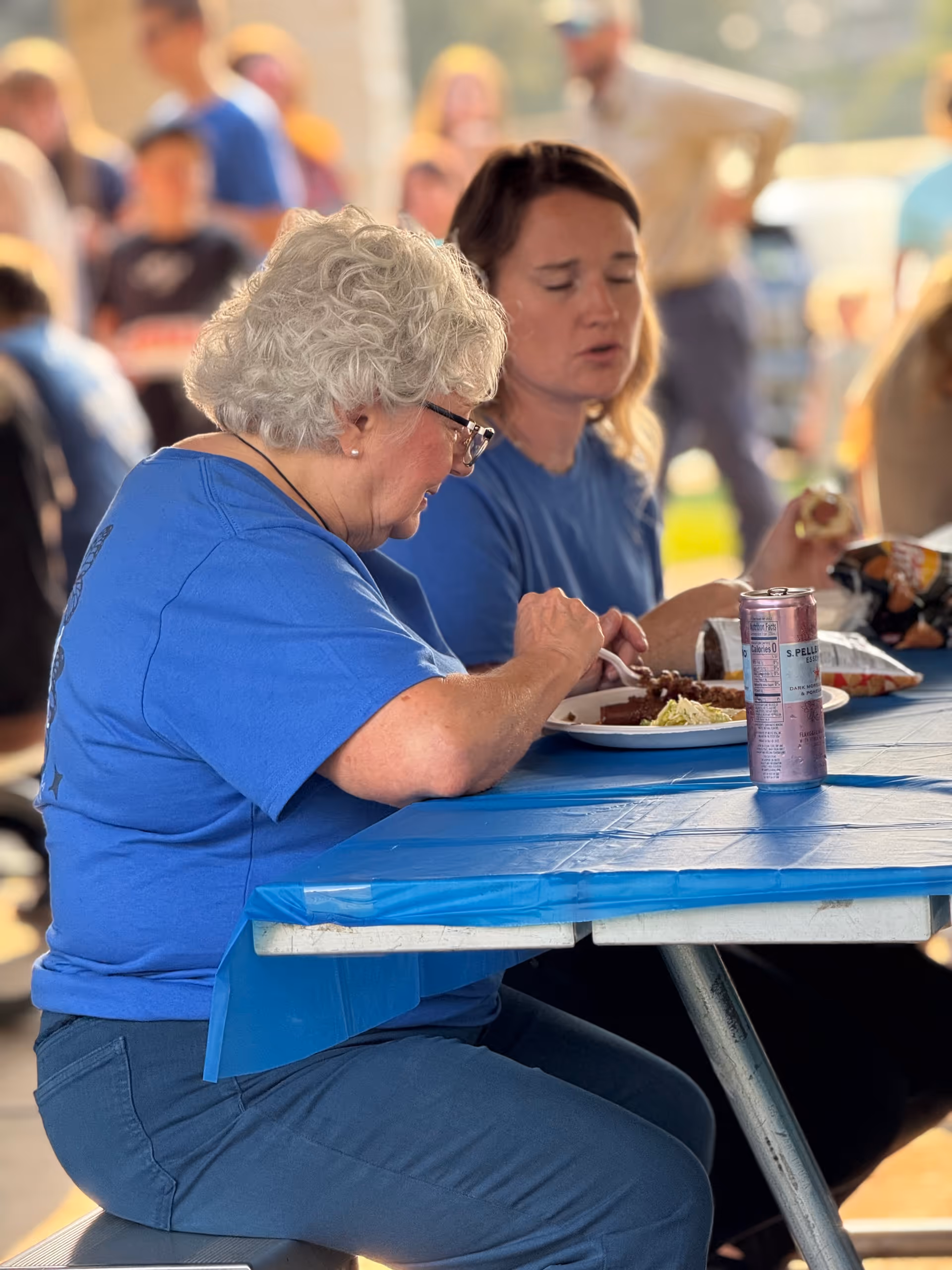 Two elderly women in blue shirts eat at a blue outdoor picnic table with others nearby.