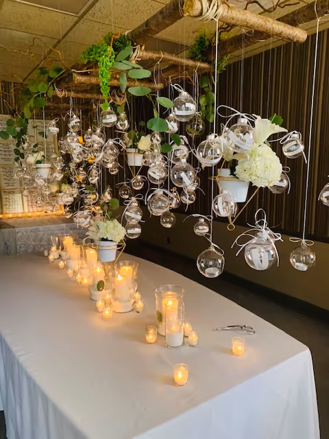 Indoor dining table covered with white cloth, candles, and hanging glass orbs with plants.