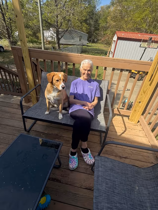 Elderly woman in a purple shirt sits on a wooden deck bench with a dog.