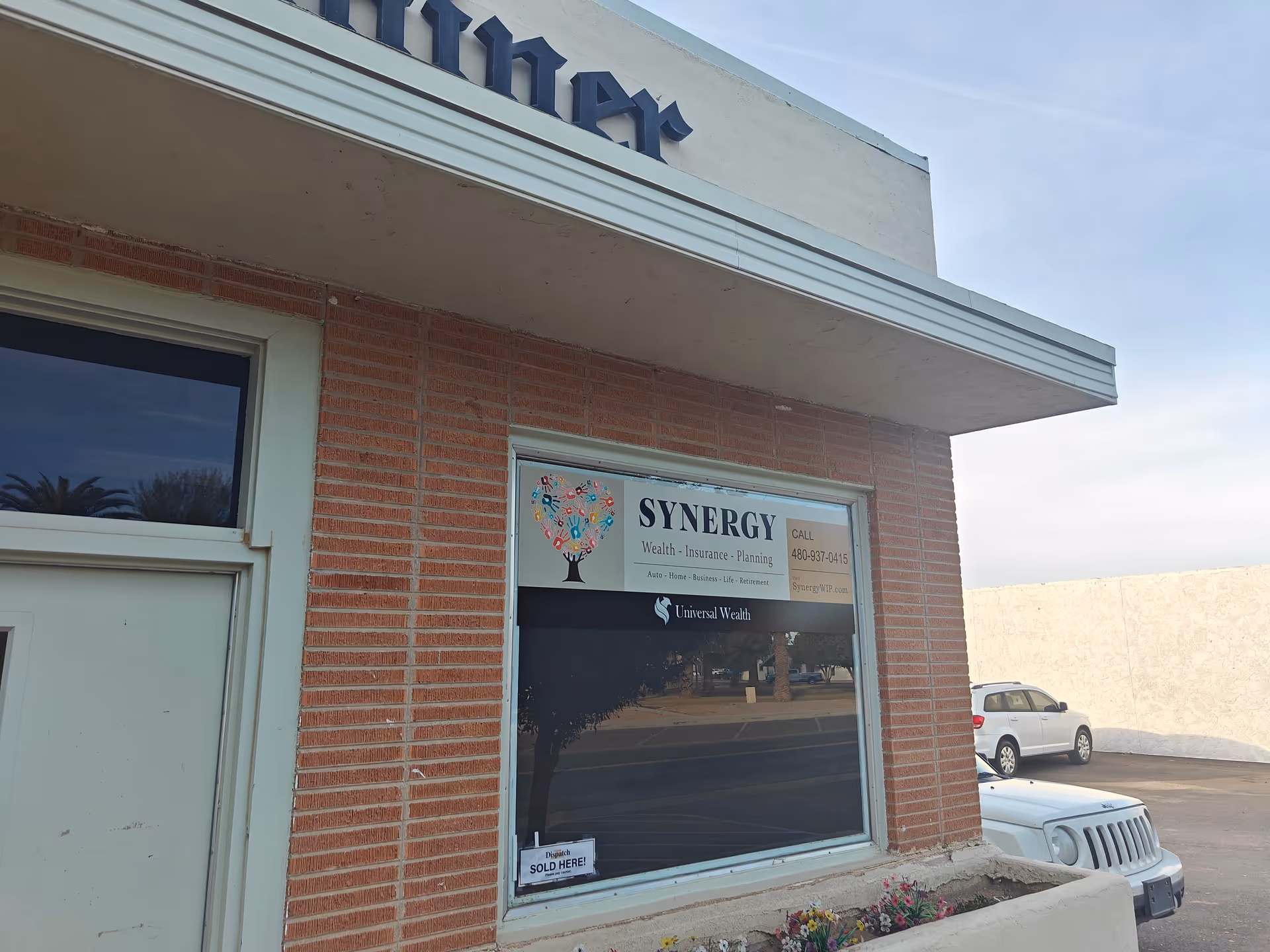 Brick building exterior with storefront window displaying Synergy Wealth sign and a parked car.