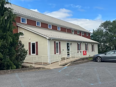 Two-story beige building with a ramp, parking lot, and trees beside it.