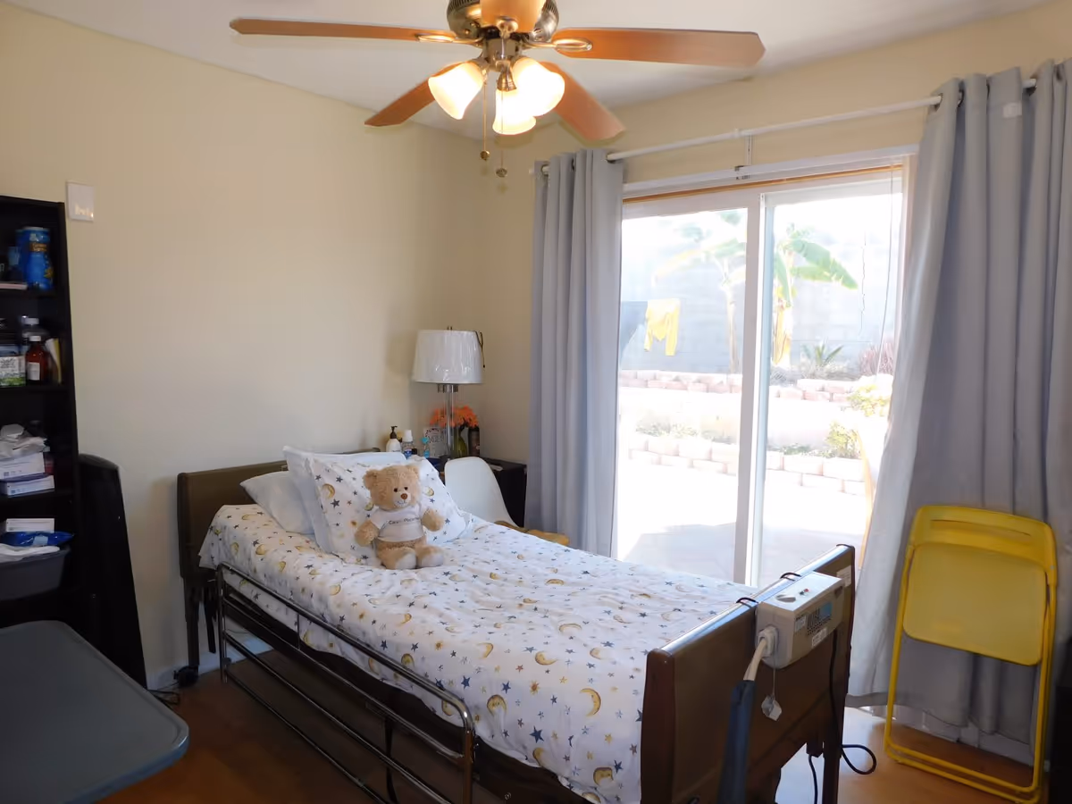 A bedroom in senior living facility with a single bed, teddy bear, and sliding glass door.