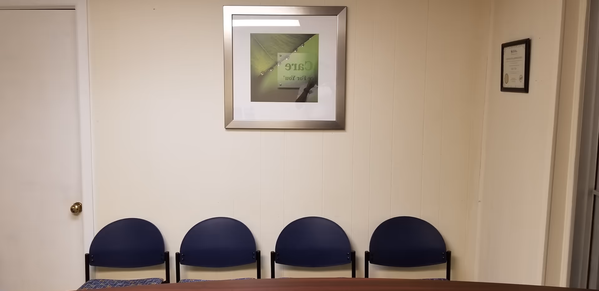 Row of blue chairs along a beige wall with framed art and a doorway.