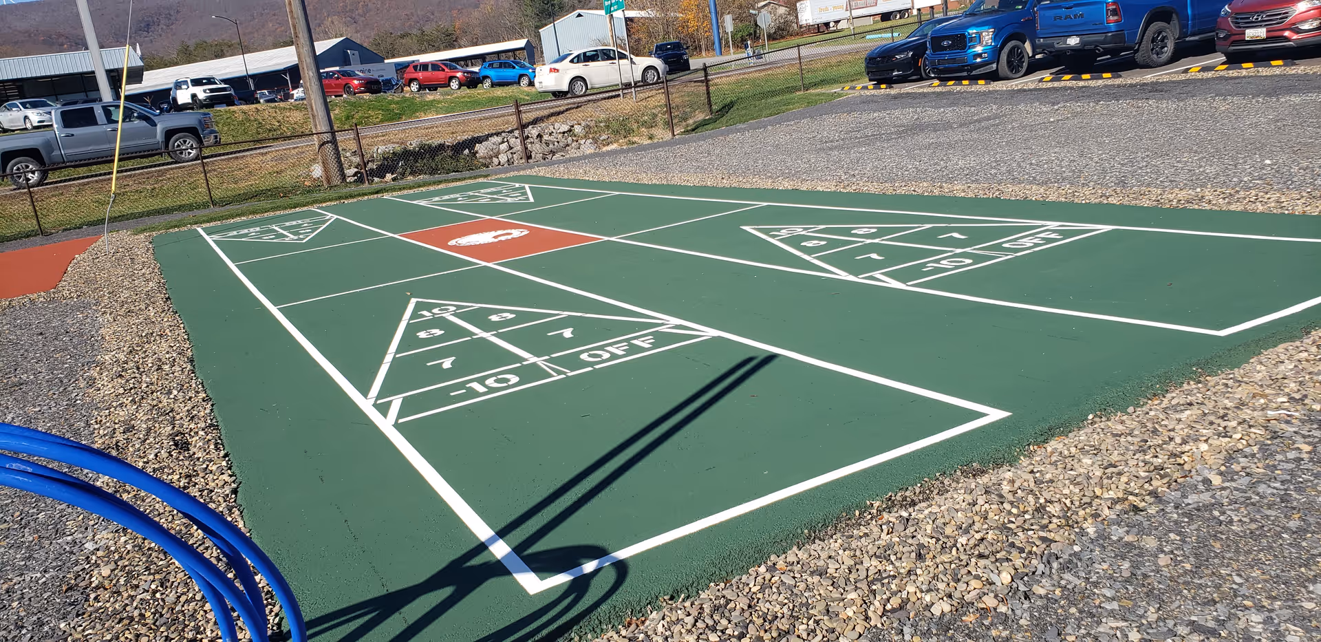 Outdoor activity court with green surface, white painted lines, gravel surrounding, and parked cars nearby.