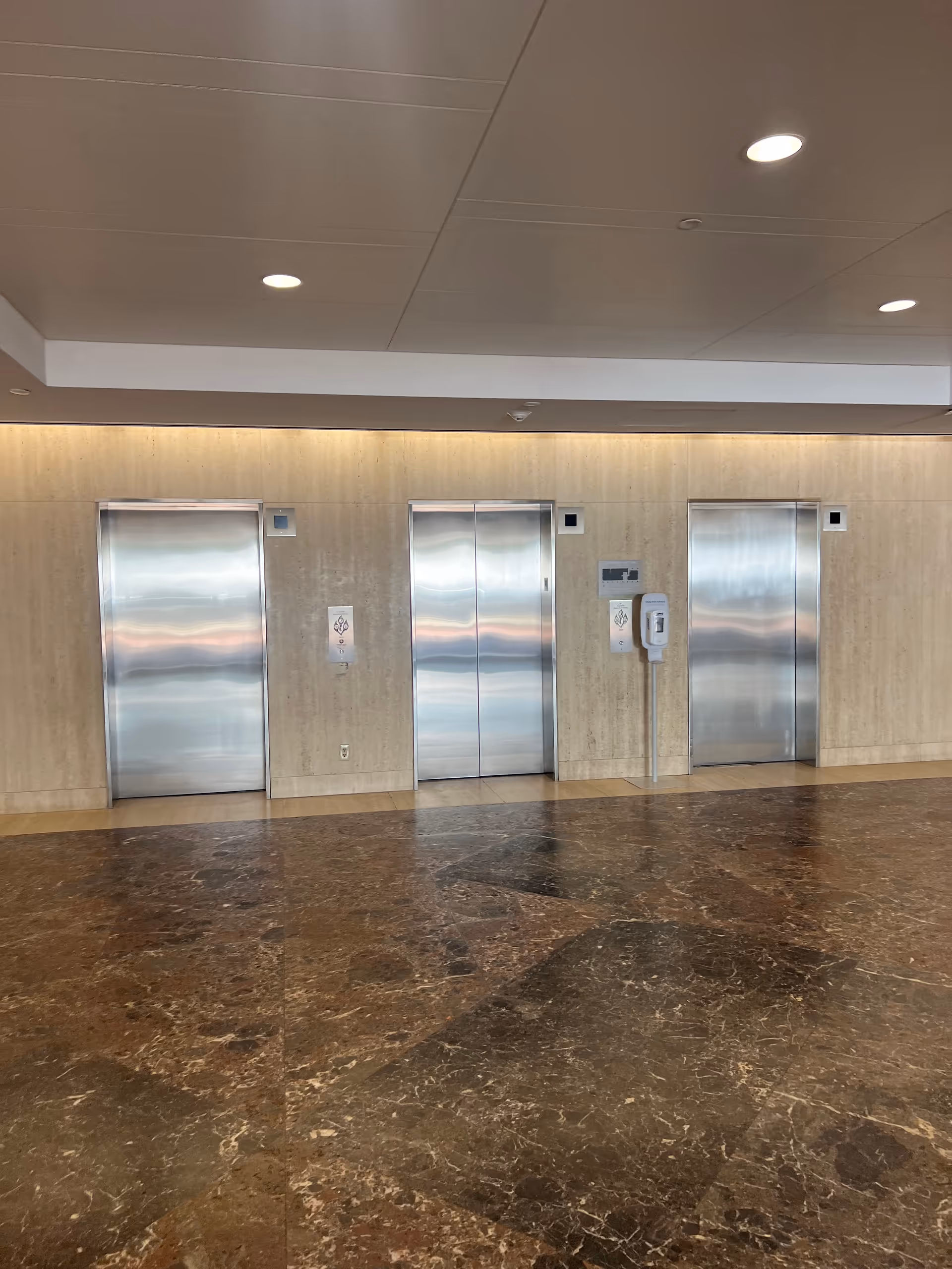 Interior hallway with three stainless elevators against a beige wall and marble floor.