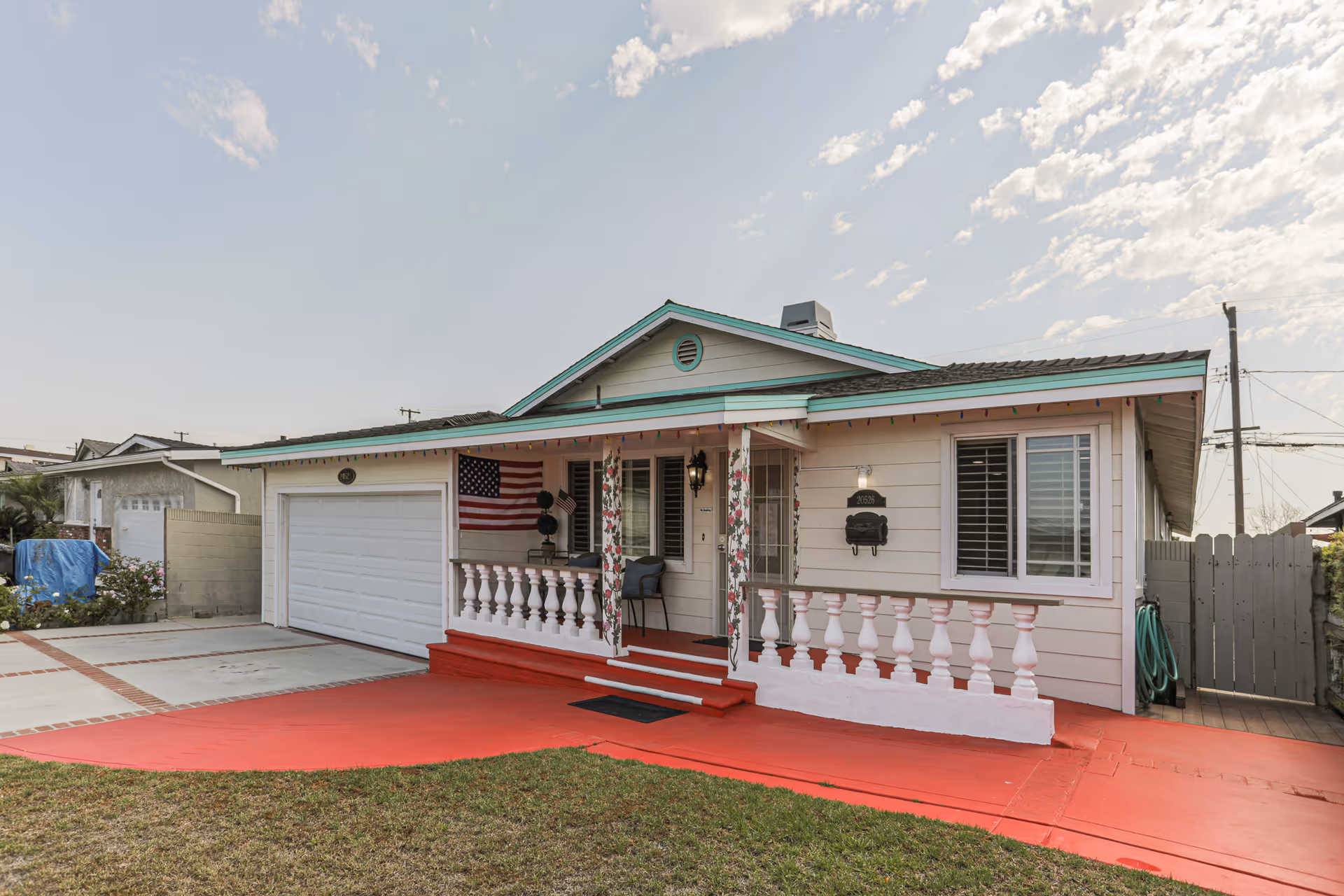 Single-story pastel house with white railings, front porch, American flag, red walkway, and garage.