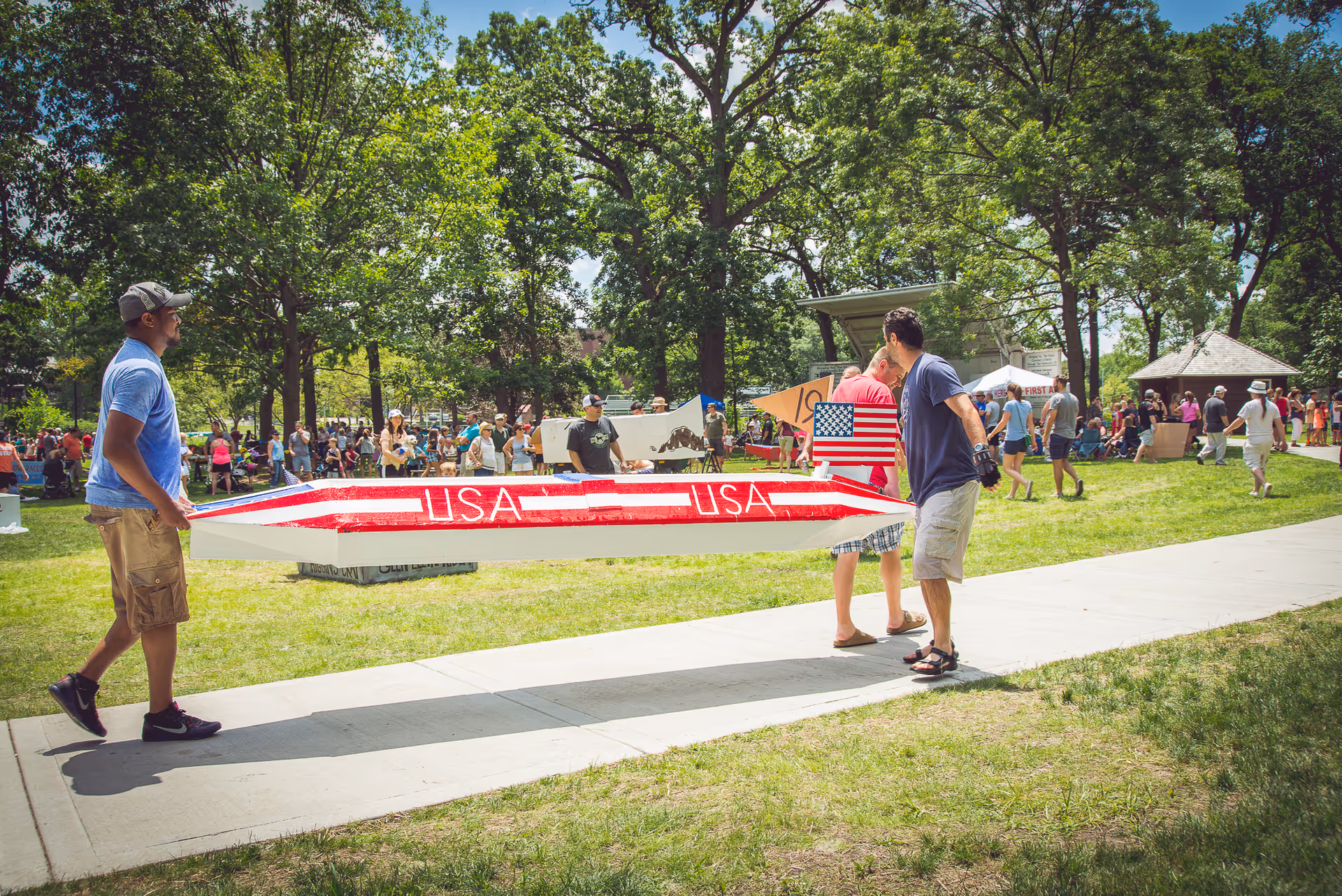 Two men carry a red, white, and blue USA canoe at an outdoor park.