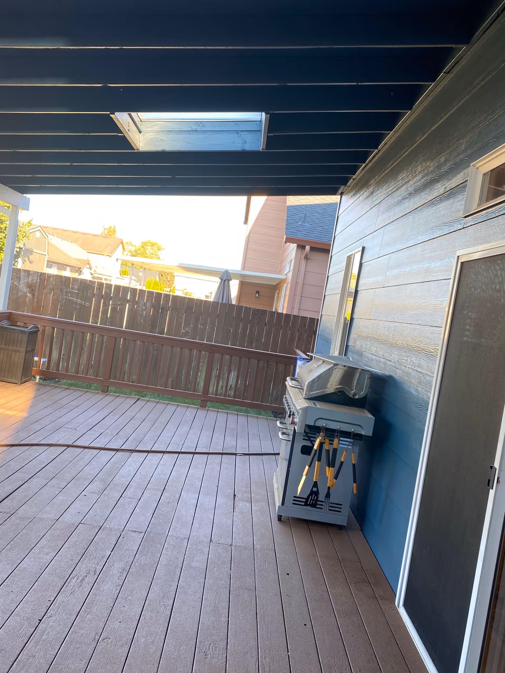 Covered wooden deck with grill and utensils beside a blue siding home.
