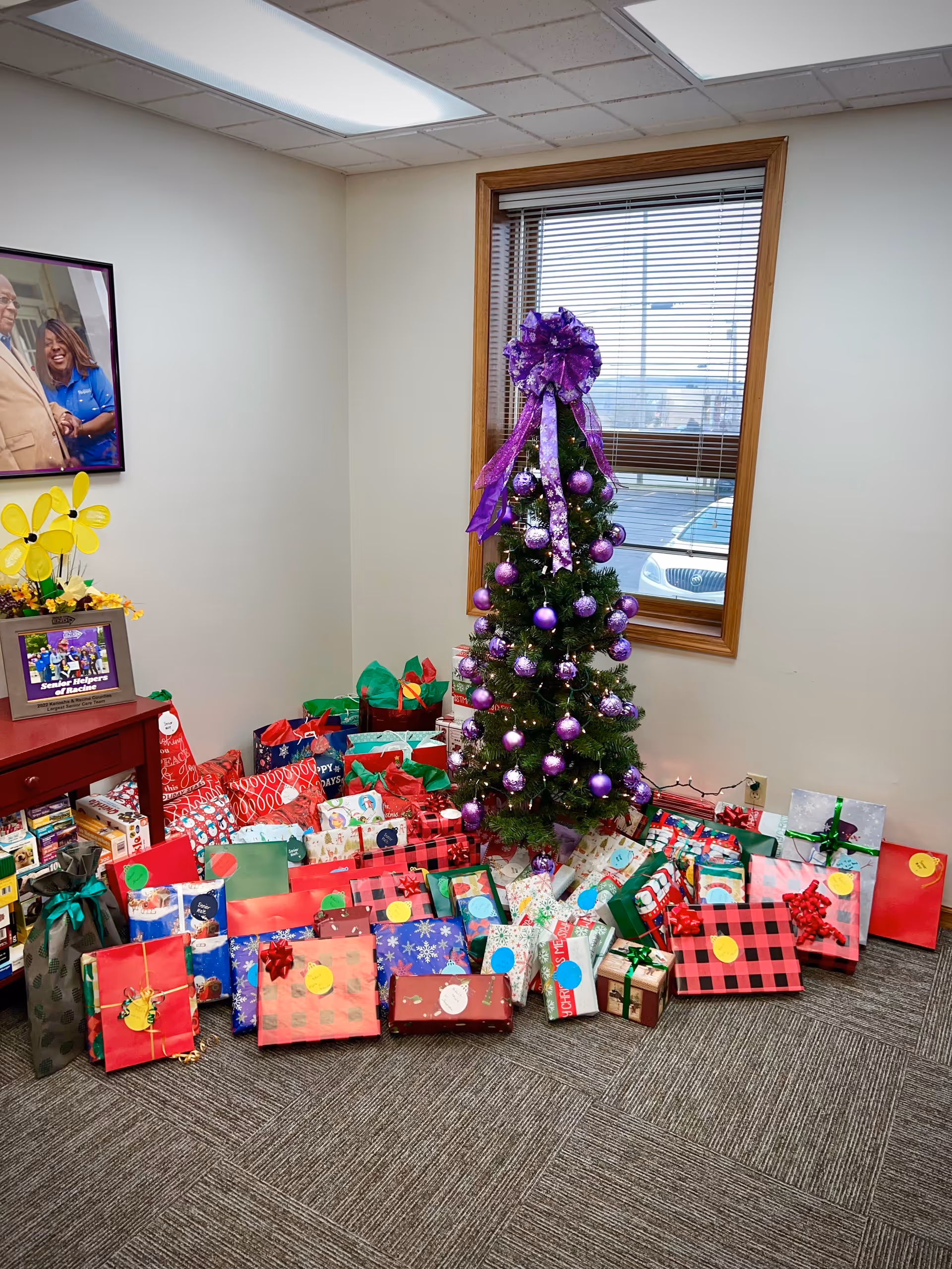 A decorated Christmas tree with wrapped gifts sits in a small office-like room.