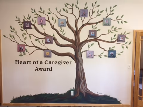 Interior wall mural of a tree titled Heart of a Caregiver Award with photo leaves.