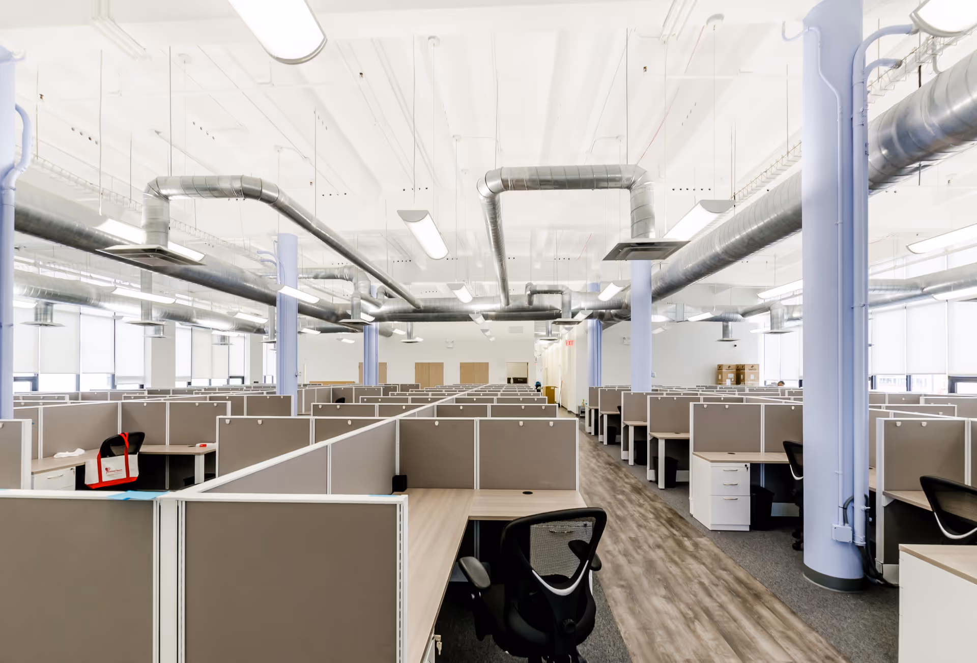 A large open office with numerous gray cubicles, desks, and exposed ductwork.