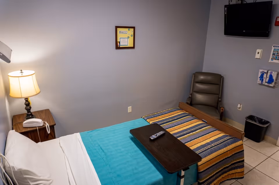 A two-bed patient room with a nightstand, lamp, chair, and wall TV.