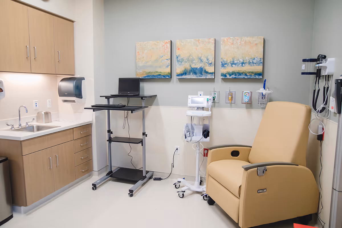 A medical examination room with a reclining chair, IV pole, medical cart, and sink.