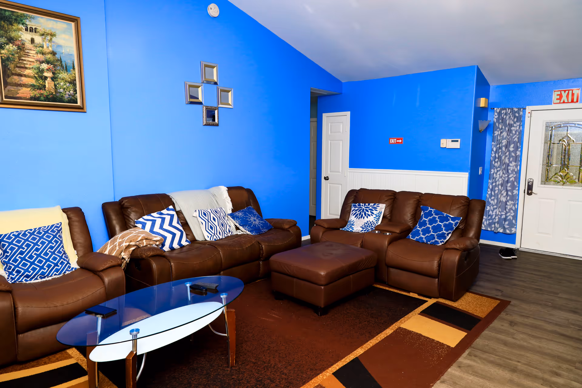 Blue-walled living room with brown leather sofas, cushions, glass coffee table, and exit doorway.