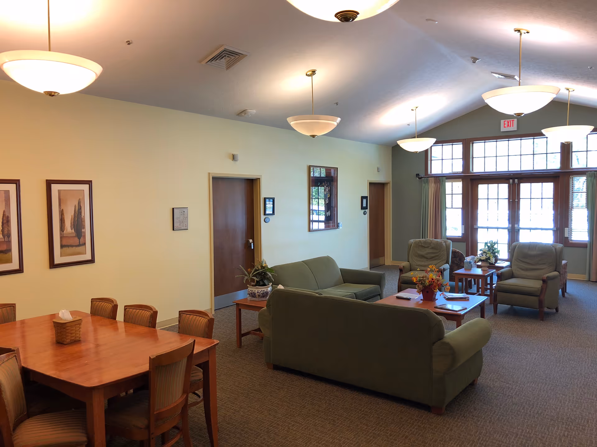 Spacious senior living lounge with green seating, wooden dining table, and large windows.