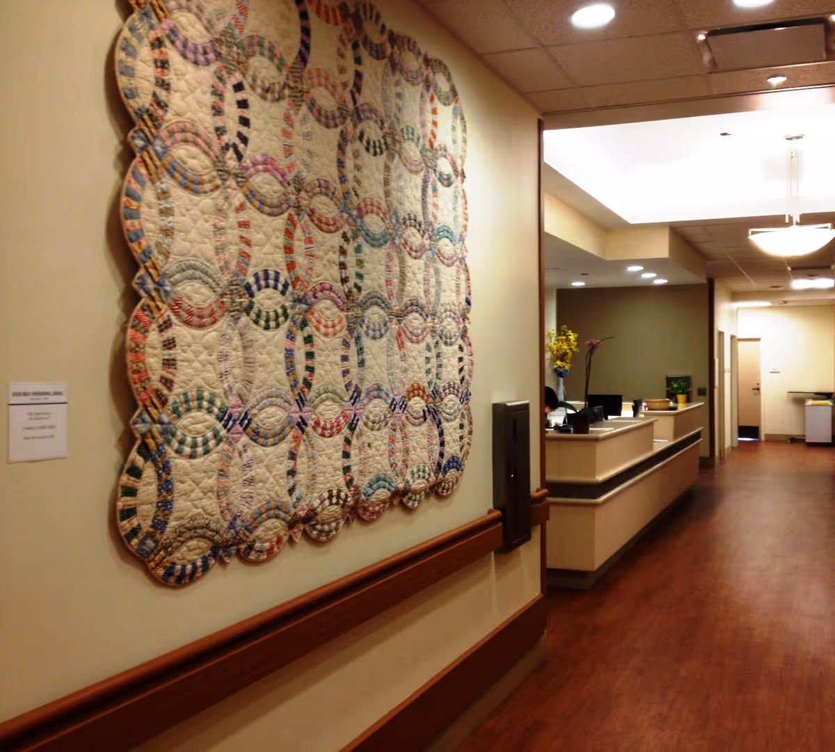 Interior hallway of a senior living facility with a large quilt on the wall near reception