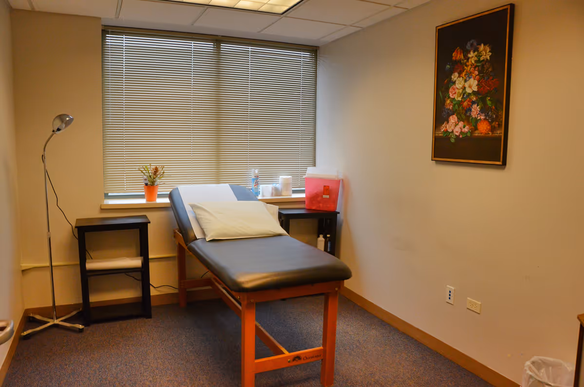 A small examination room with a padded chair, window blinds, and a floral wall painting.