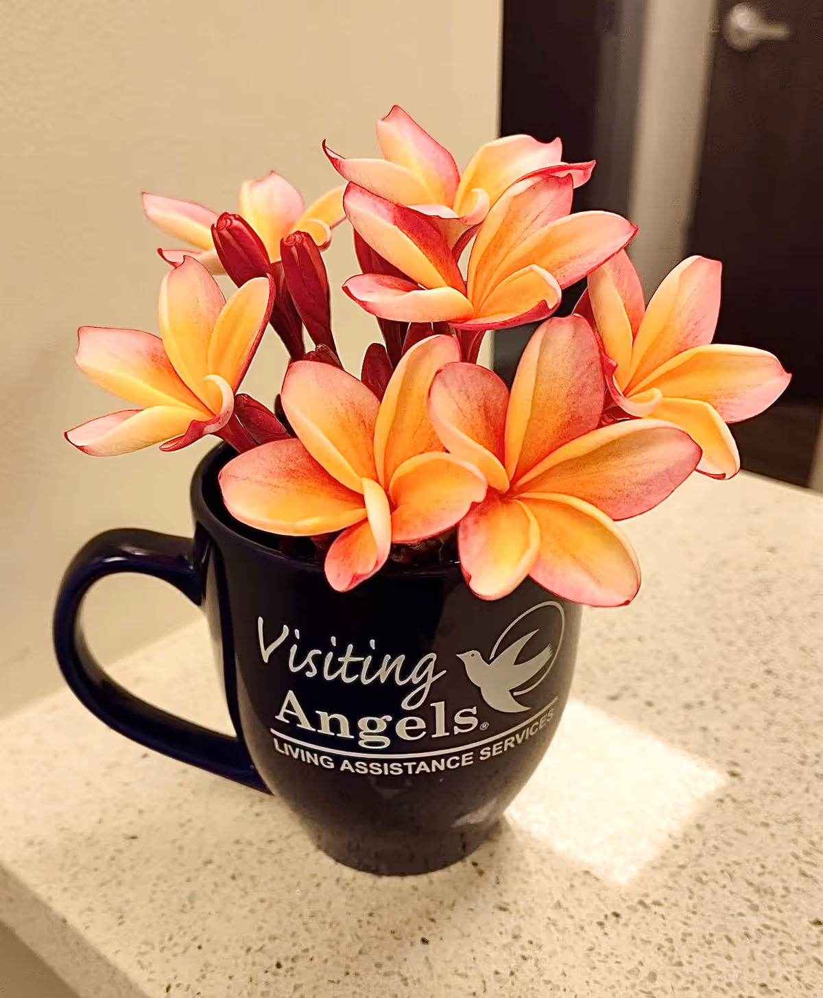 Black mug with Visiting Angels logo and orange plumeria flowers on a countertop.
