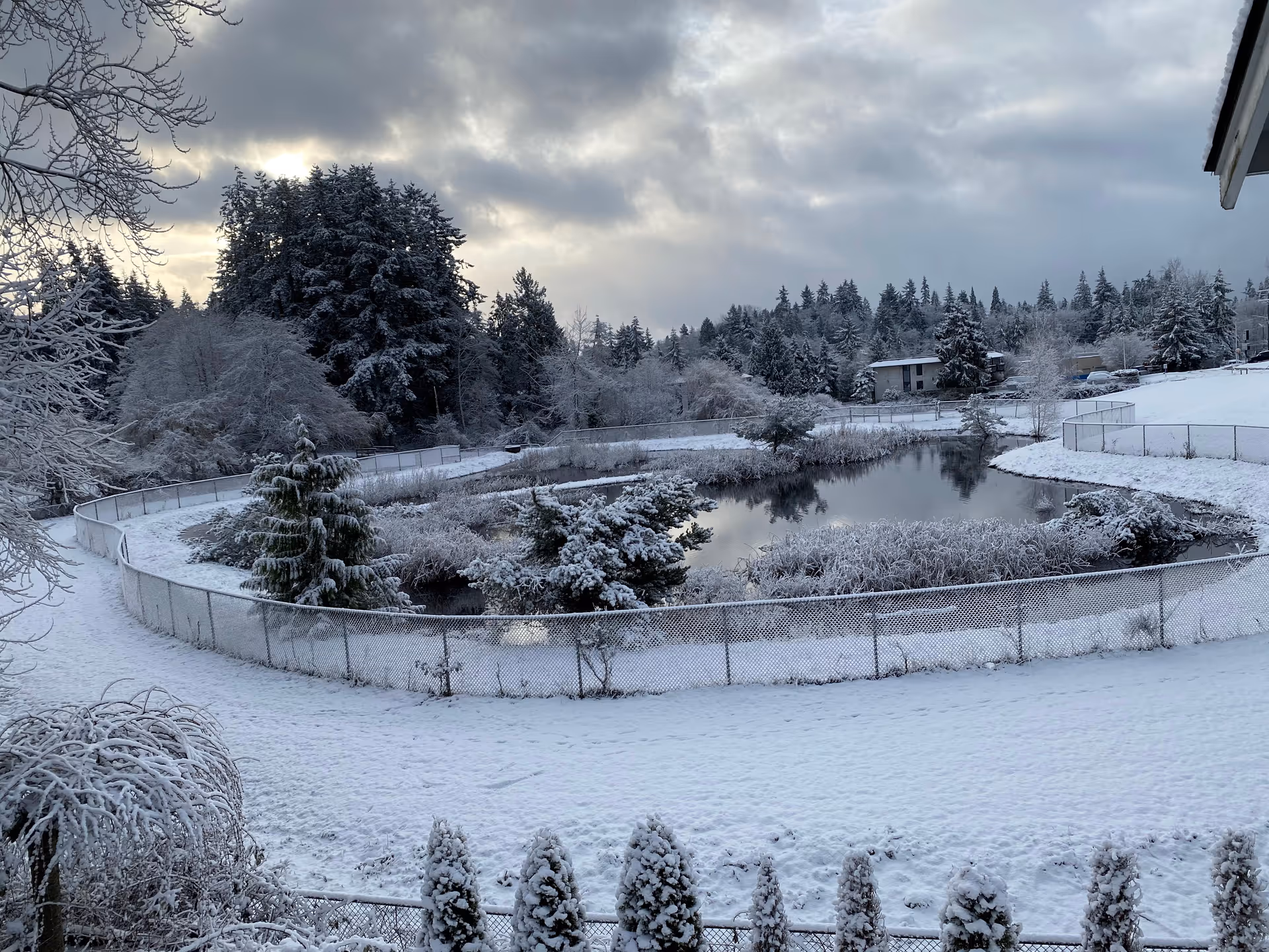 Snow-covered outdoor facility scene with a fenced pond, leafless trees, and snow-blanketed grounds.