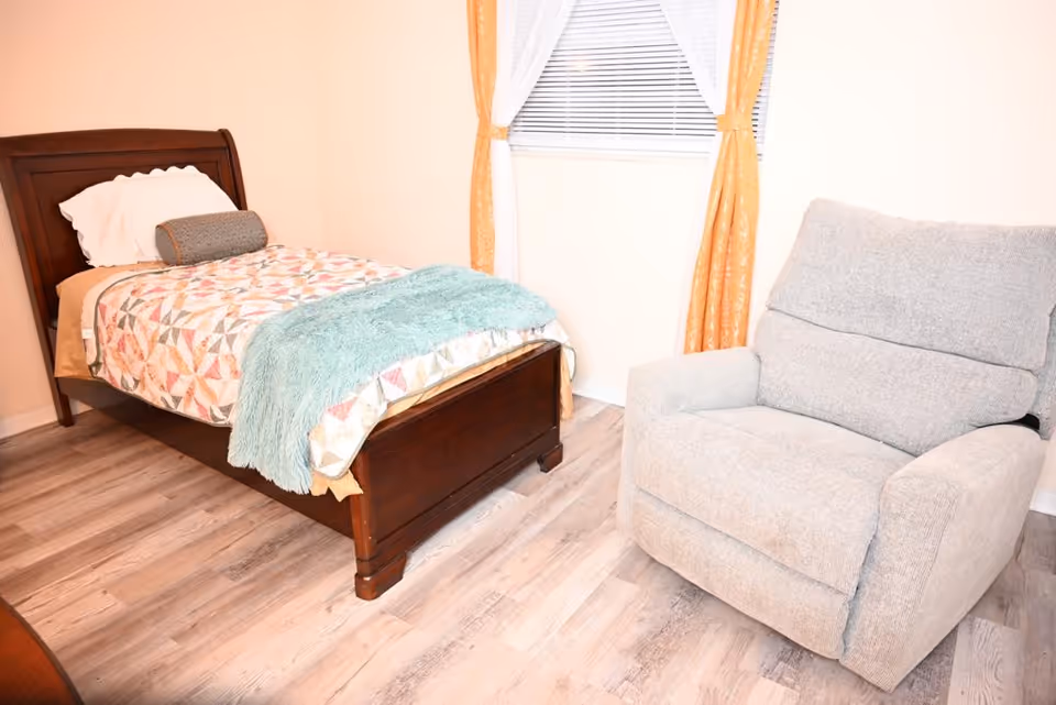 Cozy bedroom in a senior facility with a wooden bed, quilt, chair, and window curtains.