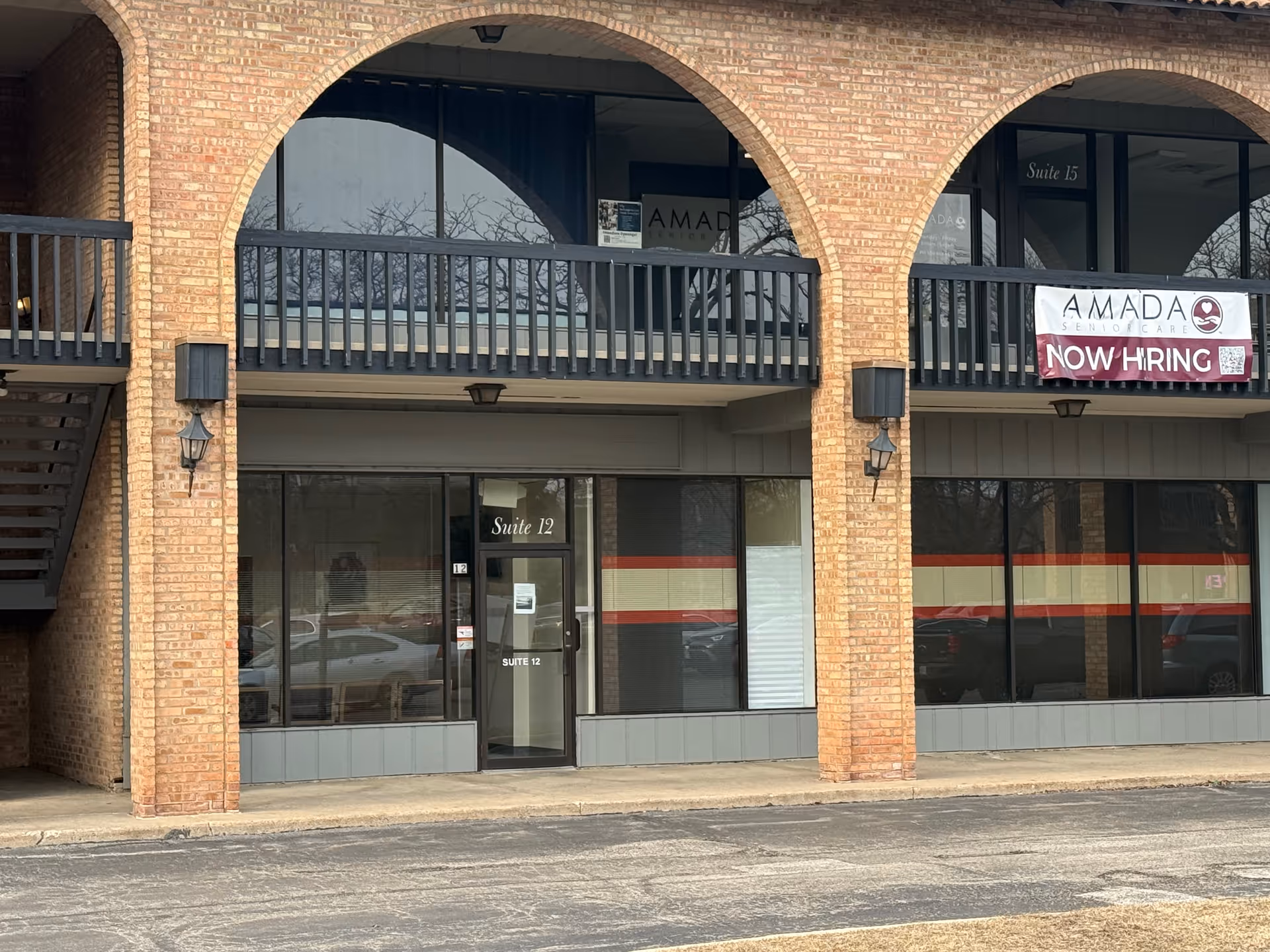 Exterior view of a brick Amada Senior Care building with arches and a Now Hiring banner.