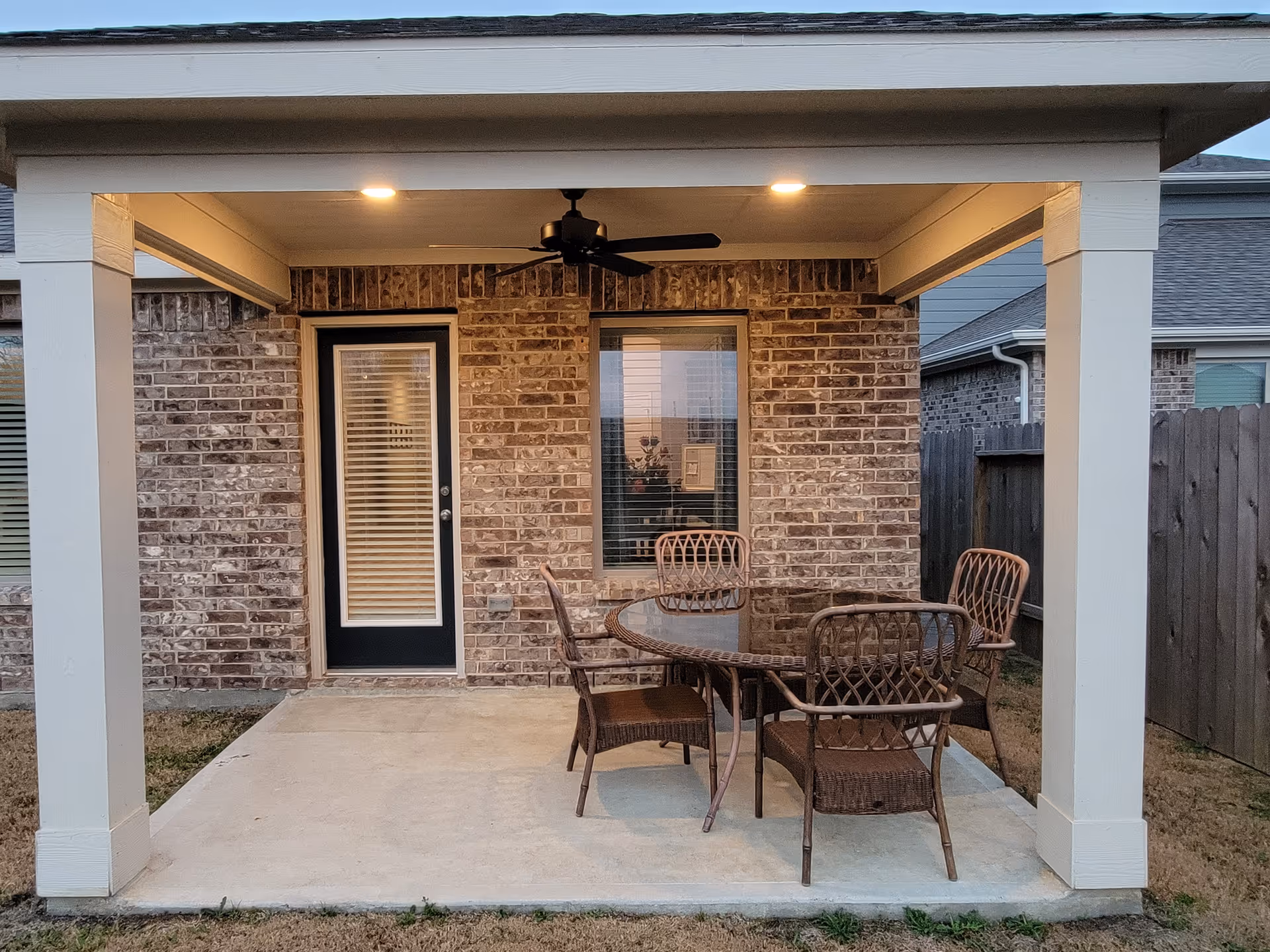 Covered brick patio with a glass door, window, ceiling fan, and wicker dining set.